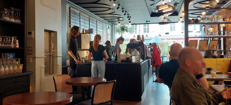 the view from the back of the cafe, looking towards the counter and the front window. There is a clear path between tables. the floor is wood and smooth, with no obstacles.