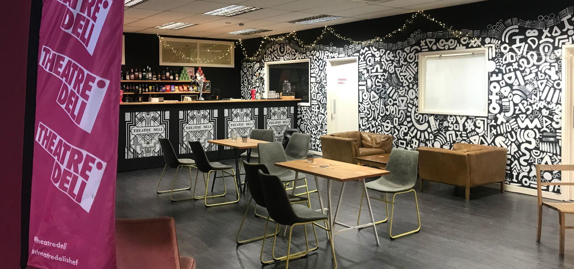 Theatre Deli's bar area with two tables, surrounded by chairs and two arm chairs next to a wall. There is a vibrant black and white mural and fairy lights, along with a flag with the company's logo and social media handles in the foreground.