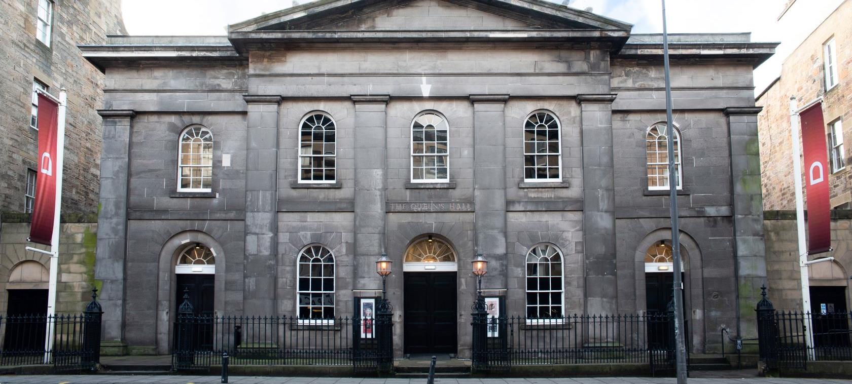The outside of The Queen's Hall is a large Georgian building with a clock tower, black doors and railings, arched windows and red flags with white arches on white flagpoles.