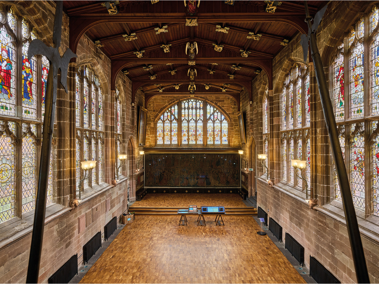 An image looking over the Great Hall at St Mary's Guildhall. In the foreground are two pikes framing the image. The wooden floor is shiny and of several different shades of colour. The stone walls have black radiators on them at ground level. There are colourful stained and painted glass windows in each wall. The wooden roof has gilded carved roof bosses, in the shape of angels with musical instruments and animals. In the centre is the Coventry Tapestry, which is quite dark in its case and cannot clearly be seen. The photographer is Andrew Moore.